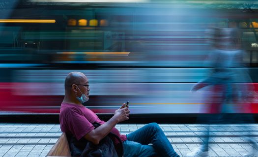 A long exposure photograph of a man sitting at a transport stop, as a vehicle goes by in background, blurred. The man is on his phone, and has a face mask pulled under his chin.